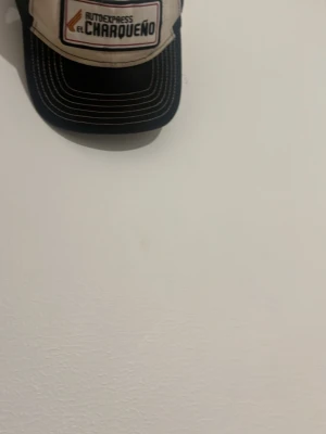Beige och svart truckerkeps El Charqueño - Säljer en beige och svart truckerkeps med broderad patch framtill där det står 'Autotransportes El Charqueño'. Kepsen har svart böjd skärm och mesh baktill för extra ventilation. Perfekt för dig som gillar retrostil och streetwear.