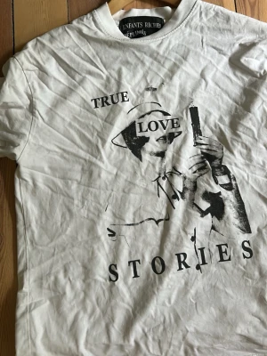 Vit t-shirt Enfants Riches Déprimés - Vit t-shirt från Enfants Riches Déprimés med svart grafiskt tryck och texten 'True Love Stories' på framsidan. Klassisk rund hals och korta ärmar. Materialet är bomull och passformen är normal. Perfekt för dig som gillar edgy och unik design.