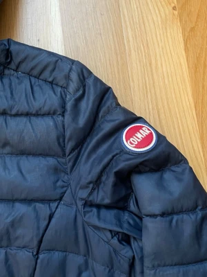 Svart dunjacka med huva från Colmar - Snygg svart dunjacka från Colmar med huva och dragkedja framtill. Jackan har två snedställda fickor med dragkedja och ett klassiskt Colmar-märke på ärmen. Perfekt för kalla vinterdagar och riktigt lätt att bära. Materialet är glansigt och quiltat.