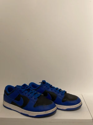 Nike Dunk Low blå och svarta sneakers - Snygga Nike Dunk Low sneakers i blått och svart läder med klassisk swoosh på sidan. Skorna har en låg siluett, matchande blå snören och vit sula med blå detaljer. Perfekta för dig som vill ha en streetwear-look med edge. Box finns men är inte i bästa kondition. Pris kan diskuteras 