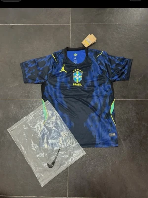 Blå Brasilien matchtröja Nike - Snygg blå och svart Brasilien matchtröja från Nike med gula detaljer och landslagets emblem på bröstet. Tröjan har korta ärmar, rund hals och är tillverkad i ett lätt, ventilerande funktionsmaterial. Perfekt för fotbollsträning eller att bära till vardags.