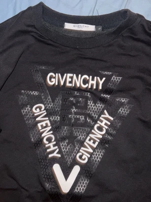 Svart sweatshirt från Givenchy - Snygg svart sweatshirt från Givenchy med stort grafiskt tryck och logga framtill. Tröjan har rund halsringning, långa ärmar och ribbade muddar vid ärmslut och nederkant. Perfekt för dig som gillar streetwear och exklusiva märken. Liten i storlek