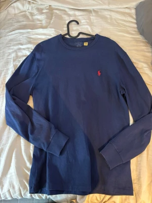 Mörkblå långärmad tröja Polo Ralph Lauren - Snygg mörkblå långärmad tröja från Polo Ralph Lauren med klassisk rund hals och röd broderad logga på bröstet. Tillverkad i mjuk bomull och har ribbade muddar vid ärmslut och hals. Perfekt för en clean och stilren look.