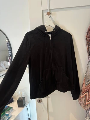 Svart velour hoodie med dragkedja -  Skön zip svart hoodie i super mjuk velour med dragkedja framtill och kängurufickor. Hoodien har långa ärmar och en klassisk huva med snörning. 