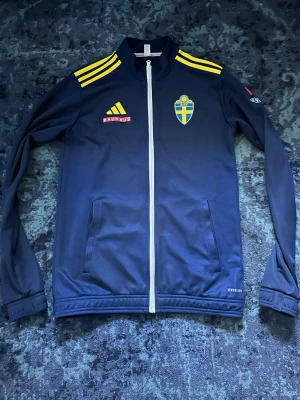 Blå Adidas Sverige träningsjacka - Mörkblå träningsjacka från Adidas med Sveriges landslagsemblem på bröstet, gula detaljer och Bauhaus-logga. Jackan har hel dragkedja, två fickor och gula ränder på axlarna. Materialet är troligtvis polyester, perfekt för sport och träning.