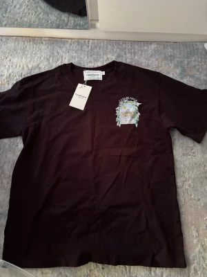 Svart t-shirt från Casablanca - Snygg svart t-shirt från Casablanca med tryck på bröstet som föreställer en arkad och palmer. Klassisk rund hals och korta ärmar. Perfekt för dig som gillar stilrena plagg med en twist. Materialet känns mjukt och skönt mot huden.