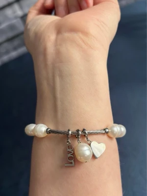 Honora Pärlarmband med Berlocker - Snyggt armband med vita pärlor. Armbandet har tre berlocker: ett hjärta, en pärla och en bricka med texten 'Love'. Perfekt accessoar för att lyfta din stil med en romantisk touch. I fint skick, stretchig.