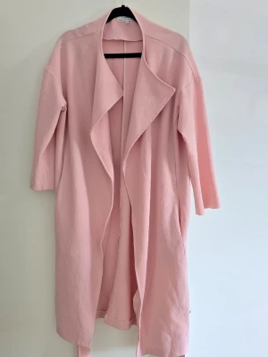 Ljusrosa trenchcoat från Closet storlek M/L - Trendig ljusrosa trenchcoat från Closet med avslappnad passform och knytskärp i midjan. Jackan har breda slag, långa ärmar och är tillverkad i ett mjukt, strukturerat tyg. Perfekt för dig som vill ha en stilren och modern look.