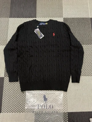 Svart kabelstickad tröja Polo Ralph Lauren - Finns I S-M-L Kabelstickad svart tröja från Polo Ralph Lauren med röd broderad logga på bröstet. Tröjan har rund halsringning, långa ärmar och ribbade muddar vid ärmslut och nederkant. Klassisk och stilren design som passar till mycket. Skriv vid intresse 