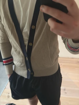 Beige kofta med marinblå detaljer - Snygg beige kofta med marinblå kant och guldknappar. Ärmsluten har röda och vita ränder som ger en sportig touch. Perfekt för lager-på-lager och passar till både jeans och shorts. Mjuk och tunn i materialet, riktigt skön att ha på sig.
