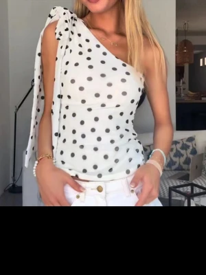 Prickig oneshoulder topp med rosett - Snygg vit oneshoulder topp med svarta prickar och stor rosett på axeln. Toppen är ärmlös och har en figurnära passform. Perfekt för dig som vill sticka ut lite extra. Materialet känns lätt och luftigt.