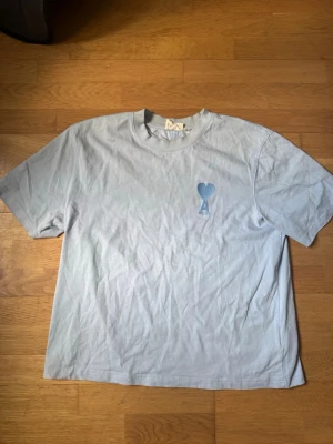 Ljusblå t-shirt från Ami Paris - Säljer en ljusblå t-shirt från Ami Paris med det ikoniska hjärta-A broderiet på bröstet. T-shirten har klassisk passform, rund halsringning och är tillverkad i mjuk bomull. Perfekt för en clean och stilren look.