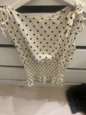 Prickig ärmlös topp med knyt - Supersöt vit topp med svarta prickar och smockad midja. Toppen är ärmlös med knytband på axlarna för en extra gullig touch. Perfekt för dig som gillar en feminin och trendig stil.