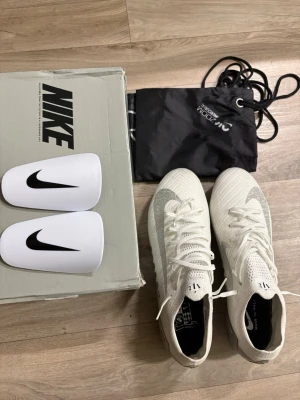 Nike vita fotbollsskor med silverdetaljer - Säljer ett par vita Nike fotbollsskor med silverdetaljer och mönstrad sula. Skorna har snörning, högt skaft och är tillverkade i syntetmaterial. Medföljer benskydd, skopåse och originalkartong. Perfekta för dig som vill sticka ut på planen. Skorna är helt nya. Priset kan diskuteras vid snabb affär