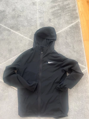 Svart Nike vindjacka med huva - Snygg svart vindjacka från Nike med dragkedja framtill och huva. Jackan har en stilren design med Nike-logga på bröstet och är tillverkad i ett lätt material som skyddar mot vind. Perfekt för en sportig och avslappnad look.
