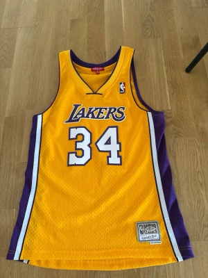 Lakers basketlinne #34 Mitchell & Ness - Gult och lila basketlinne från Mitchell & Ness med Lakers-tryck och nummer 34. Klassisk NBA-design med broderade detaljer och meshmaterial som andas. Perfekt för basketträning eller som streetwear. 
