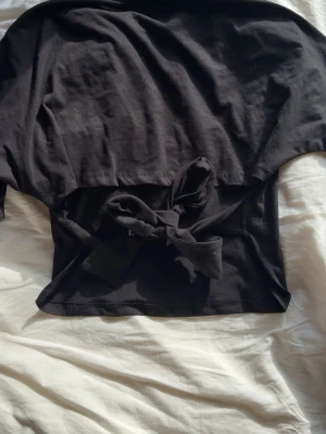 Svart topp med knyt i ryggen  - Svart topp med rund halsringning och vida ärmar. Toppen har en snygg knytning i midjan som ger en cool och trendig look. Tillverkad i mjuk bomull, perfekt för att lyfta din outfit med en enkel men stilren detalj.