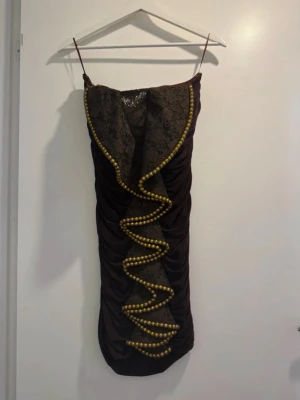 Brun spetsklänning med gulddetaljer - Unik brun klänning med smala axelband, draperad passform och dekorativ spets framtill. Klänningen har en vågig kant prydd med guldiga pärlor som ger en cool och iögonfallande look. Perfekt för dig som vill sticka ut med en annorlunda stil.