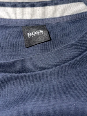 Mörkblå kofta från Hugo Boss - Snygg marinblå kofta från Hugo Boss med stort vitt BOSS-tryck på ryggen. Klassisk modell med ribbade muddar och mjukt material som känns skönt mot huden. Perfekt för dig som gillar stilrena och trendiga plagg med tydlig logga.