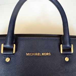 Marinblå handväska från Michael Kors - Skitsnygg mörkblå Michael Kors väska Perfekt storlek till skola och jobb  Ganska säker på att det är modellen Selma, Medium Leather Satchel i färgen marinblå  Köpt i en fin vintage butik där den såldes som en äkta Michael Kors men vågar inte lova något. Den är fint gjord, bra kvalitet och alla graveringar och märkningar ser äkta ut! Hur som helst är den rejäl och bra i kvaliteten och i toppen skick, en riktig fin väska.  Skriv gärna om ni har några andra frågor! 