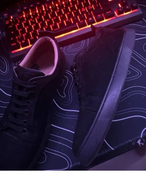 Svarta sneakers i mocka-look - Snygga svarta sneakers med klassisk design och låga skaft. Skorna har svarta snören och en svart sula, samt ovandel i mocka-liknande material. Perfekta för en stilren och clean streetwear-look.