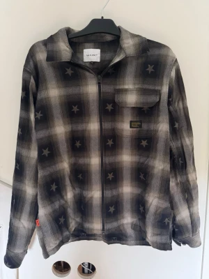 Svart och grå overshirt från Valient - Overshirt från Valient i svart och grå rutigt mönster med stjärnor. Modellen har dragkedja framtill, en bröstficka med lock och klassisk krage. Materialet är mjukt och passar perfekt för lager-på-lager. Snygg detalj med patch på bröstet.