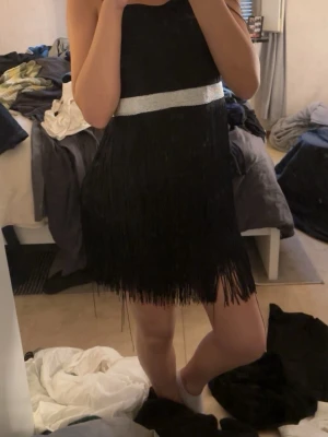 Svart fransklänning med silverdetalj - Snygg svart kortklänning med fransar och en bred silverfärgad detalj i midjan. Klänningen är ärmlös och har en rak passform som ger en cool och festlig look. Perfekt för dig som vill sticka ut med stilrena detaljer och mycket rörelse i tyget.