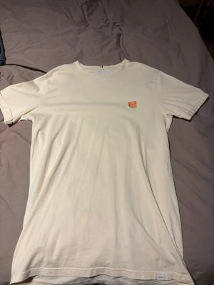 Vit t-shirt från Les Deux - Vit t-shirt från Les Deux med korta ärmar och rund hals. På bröstet sitter en liten orange patch med vit brodyr. T-shirten har en clean och stilren look, perfekt till jeans eller shorts.