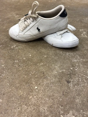 Vita Polo Ralph Lauren sneakers - Snygga vita sneakers från Polo Ralph Lauren med klassisk låg profil och svart detalj vid hälen där det står 'POLO'. Skorna har den ikoniska hästloggan på sidan och är tillverkade i läder med vita snören och gummisula. Perfekta för en clean och stilren look.