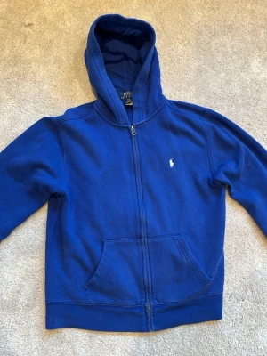 Blå hoodie med dragkedja från Polo Ralph Lauren - Det som ser ut som en stor fläck på baksidan är bara Skuggan från mobilen.                                                                  Blå hoodie med dragkedja från Polo Ralph Lauren. Klassisk design med huva och vit broderad logga på bröstet. Mjuk bomullskvalitet som är skön att ha på sig. Perfekt för en chill och avslappnad stil.