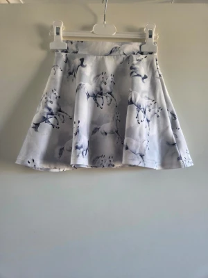 Vit kjol med hästmönster, 98/104 - Superfin vit kortkjol med grå hästmönster. Kjolen har en utsvängd form och är tillverkad i ett mjukt och bekvämt material, perfekt för dig som gillar unika prints och vill sticka ut lite extra.