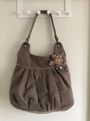 Beige handväska med tygblomma - Snygg beige handväska i tyg med veckad design och axelrem. Väskan har dekorativa paljetter och en stor tygblomma i dova färger samt två glittriga broscher framtill. Perfekt accessoar för att lyfta din outfit.