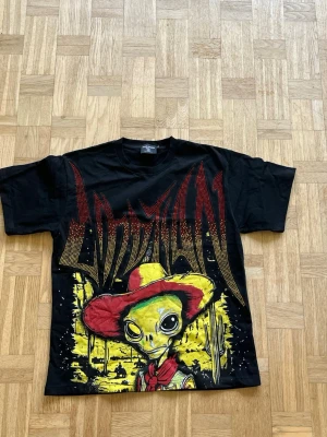 Mixed Emotions T-Shirt - Cool svart t-shirt med stor färgglad print av en alien i röd cowboyhatt och scarf på framsidan. Bakstycket har tryck med UFO och grafiska detaljer i rött och gult. T-shirten har normal passform och korta ärmar. 