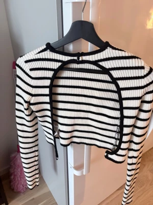 Randig croppad bolerotopp - Snygg croppad bolerotopp i ribbad bomull med svartvita ränder och lång ärm. Toppen har en unik öppen framdel. Perfekt för att lyfta din outfit med en cool detalj.