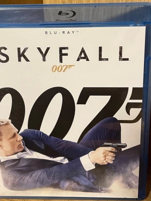Skyfall 007 Blu-ray - Skyfall 007 på Blu-ray, en actionfylld James Bond-film i klassiskt fodral av blå plast. Omslaget har en stilren design med Bond i kostym och pistol, samt tydliga 007-loggor. Perfekt för dig som gillar spänning och klassiska agentfilmer.