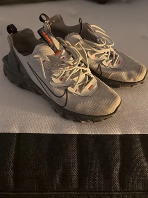 Nike React vision sneakers grå - Nike React Vision  sneakers i grått med svarta detaljer och reflexinslag. Skorna har en chunky svart sula, vita snören med röda accenter och en stor svart Swoosh på sidan. Ovandelen är i textil och syntet, perfekt för en sportig och urban stil.