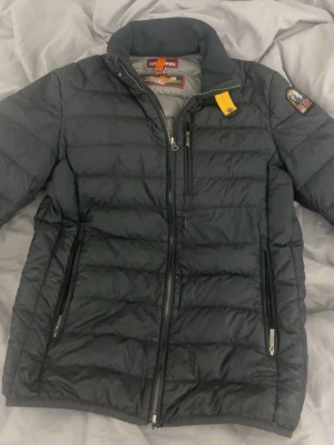 Svart dunjacka från Parajumpers - Snygg svart dunjacka från Parajumpers med klassisk gul detalj vid kragen och logotyp på ärmen. Jackan har dragkedjor på fickorna och en bröstficka med dragkedja. Perfekt för kalla vinterdagar och riktigt stilren look.