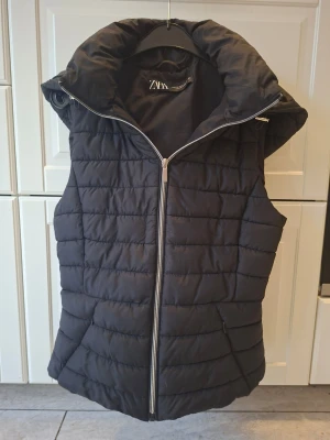 Svart lättvadderad väst från Zara - Snygg svart dunväst från Zara med quiltad design och hög krage. Västen har dragkedja framtill och är perfekt för lager-på-lager-stil. Tillverkad i lätt material som håller värmen utan att kännas klumpig. Enkel att matcha med olika outfits.