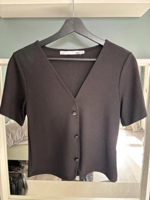 Svart topp med v-ringning och knappar - Svart topp från 157 med korta ärmar, v-ringning och tre dekorativa knappar framtill. Enkel och stilren design som passar till många olika outfits. Materialet känns mjukt och stretchigt.
