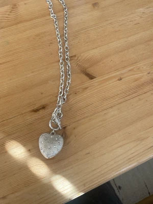 Silverfärgat halsband med hjärthänge - Stilrent halsband i silverfärg med grov kedja och ett stort hjärthänge. Hänget har en prickig och hjärtformad struktur som ger en cool och unik look. Perfekt accessoar för att lyfta din outfit.