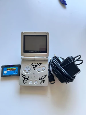 Game Boy Advance SP silver tribal - Game Boy Advance SP i silver med svart tribal-mönster. Konsolen är hopfällbar och tillverkad i plast och metall. Medföljer laddare och spelet Mario Kart: Super Circuit. Perfekt för dig som gillar retrospel och portabel gaming.