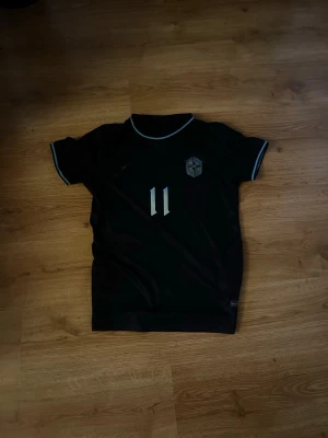 Svart Neymar Jr fotbollströja - Snygg svart fotbollströja med Neymar Jr och nummer 11 på ryggen. Tröjan har korta ärmar, ljusblå detaljer vid hals och ärmslut samt klubbmärke på bröstet. Perfekt för dig som älskar fotboll. Enda defekten är att nummret har fallit av i tvätten 