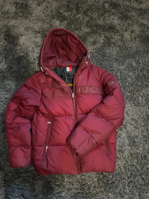 Vinröd pufferjacka från Tommy Hilfiger - Säljer en vinröd pufferjacka från Tommy Hilfiger med huva och dragkedja. Jackan har logotypen på bröstet och ett märke på ärmen. Flera praktiska fickor med dragkedja och en schysst, lite boxig passform. Perfekt för kalla dagar och riktigt snygg färg. Den är använd ett få par gånger. Inga defecter