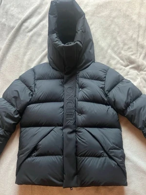 Moncler madeira  - Snygg svart dunjacka från Moncler med hög krage och stor huva. Jackan har breda, quiltade paneler och Moncler-logga på ärmen samt diskret logotyp längs dragkedjan. Perfekt för kalla vinterdagar och riktigt varm.