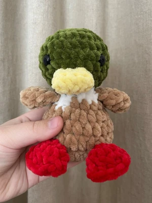 Virkad anka i naturfärger – handgjord plushie - Handvirkad anka i grönt, brunt och vitt med röda fötter. Mjuk och charmig med fina färger, perfekt som present eller inredningsdetalj. Varje anka är handgjord. Storlek: ca 15 cm.