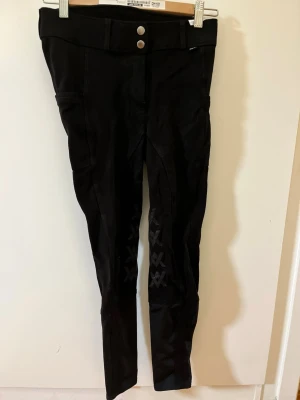 Svarta ridleggings med hög midja - Snygga svarta ridleggings med hög midja och två silverfärgade knappar framtill. Stretchigt material och diskreta detaljer på benen. Perfekta för dig som gillar en sportig och stilren look.