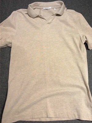 Beige polotopp från Bläck - Stilren beige polotopp från Bläck i mjukt material. Toppen har klassisk krage och korta ärmar, perfekt för en clean och avslappnad look. Passar dig som gillar enkel och tidlös stil.