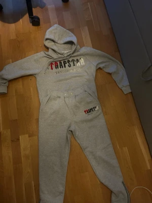 Trapstar tracksuit - Snygga gråa tracksuit från Trapstar med broderad logga i rött, svart och vitt på ena benets byxorna har resår i midjan, fickor och dragkedjor vid bensluten. Perfekta för en avslappnad och trendig stil.
