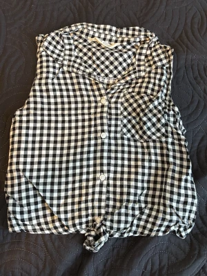 Svartvit rutig ärmlös blus - Snygg ärmlös blus i svartvitt rutigt mönster med knappar framtill och en liten bröstficka. Blusen har en knytning nertill för en avslappnad look. Perfekt till jeans eller shorts för en cool stil.