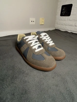 Grå och beige sneakers med gummisula - Snygga sneakers i grått och beige med vita skosnören och klassisk gummisula. Ovandelen är i mocka och läder, vilket ger en stilren och tidlös look. Perfekta för dig som gillar minimalistisk stil och vill ha något som funkar till det mesta.
